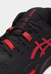 Μαύρο και κόκκινο παπούτσι τένις ASICS με έμφαση στα κορδόνια, τη γλώσσα με το λογότυπο, την υφασμάτινη επιφάνεια και το χαρακτηριστικό λογότυπο στο πλάι.