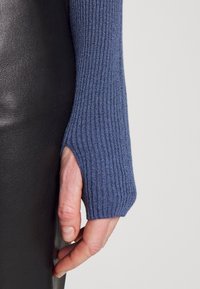 Žebrovaný pletený svetr v modré barvě s otvorem na palec. Detailní záběr ruky na černém koženém pozadí, který ukazuje texturu a design.