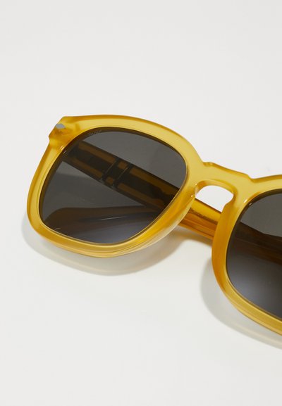 Óculos de sol de plástico amarelo com lentes escuras, apresentando uma forma geométrica arredondada e um design de ponte recortado notável.