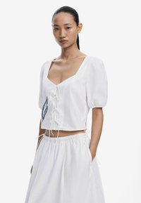 Witte cropped blouse met pofmouwen, vierkante halslijn en strikkoning aan de voorkant. Heeft blauwe blad-geborduurde details aan de linkerkant.
