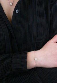 Bracciale in argento con catena, caratterizzato da un pendente circolare con una pietra centrale, indossato su un polso vicino a una camicia nera plissettata.