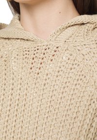 Beige stickad hoodie med en texturerad, öppen vävdesign. Har en dragsko i huvan och en avslappnad passform. Materialet verkar mjukt och lätt.