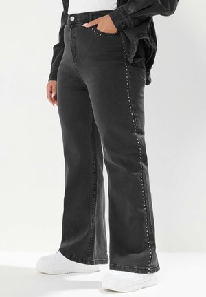 Femme portant un jean noir taille haute avec des clous sur les côtés, des baskets blanches, un haut blanc et une veste noire, debout sur un fond blanc.