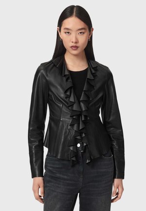 Junge Frau mit langen dunklen Haaren, die eine schwarze Lederjacke mit geraffter Vorderseite und dunkle Jeans trägt, stehend vor einem schlichten hellen Hintergrund.