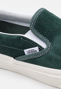 Zielone zamszowe trampki z białym elastycznym wzmocnieniem i detalami przeszycia, z białą metką logo Vans i kremową gumową podeszwą.