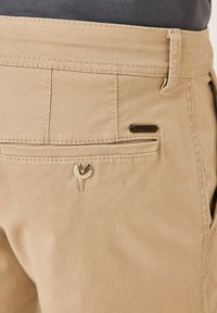 Beige cotton trousers with a textured finish, featuring a back pocket with a button closure and a small branded tag near the pocket.