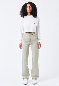Felpa bianca corta con logo piccolo, abbinata a jeans a vita alta di un verde chiaro con gamba larga e sneakers bianche. Texture del tessuto lisce.