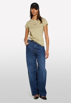 T-shirt kaki avec le logo "MUSE", jeans bleus à jambe ample et casquette noire. La tenue présente une coupe décontractée et un design casual avec des accents minimaux.