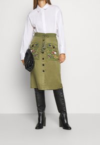 Falda midi verde oliva con bolsillos frontales, bordado decorativo y botones. Combinada con una camisa blanca y botas negras hasta la rodilla.