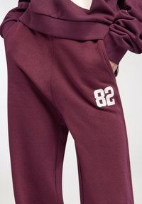 Burgundowe dresy z białym napisem "82" na udzie, zestawione z pasującą burgundową bluzą, ręce w kieszeniach.
