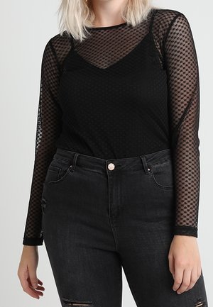 Femme portant un haut noir transparent à manches longues avec un motif losange sur un caraco noir, associé à un jean noir taille haute.