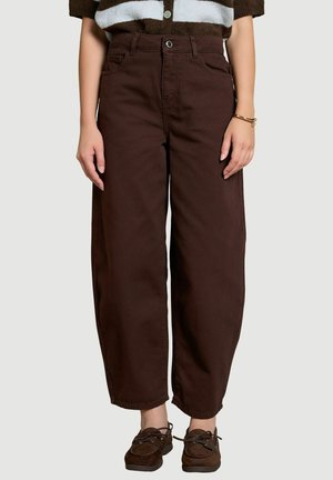 Persona que lleva pantalones marrón oscuro de talle alto y pernera ancha con bolsillos y zapatos marrones con cordones, de pie sobre un fondo claro.