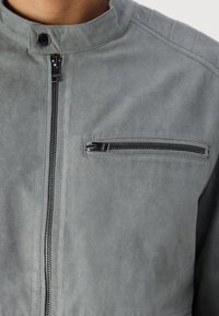 Jachetă din suede gri cu guler înalt, închidere cu fermoar în față și un mic buzunar cu fermoar pe piept. Textură moale și design minimalist.