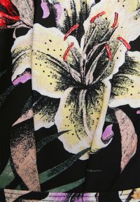 Um tecido preto apresenta um grande e detalhado lírio amarelo com estames vermelhos, rodeado por folhas verdes e suaves destaques em rosa.