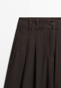 Donkerbruine rok met hoge taille, ceintuurlussen, voorste knoop, plooien en zijzakken op een witte achtergrond.
