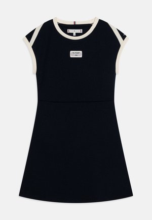 Vestido negro sin mangas en línea A con ribete crema en el cuello y las mangas, parche frontal que dice "Hilfiger 1985" y el logo de Tommy Hilfiger en la manga.