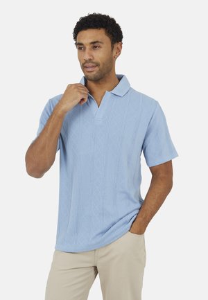 Homme aux cheveux courts et bouclés portant une chemise bleu clair à manches courtes avec col et un pantalon beige, ajustant le col avec une main.