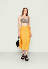 Top cropped com padrão floral e alças finas, combinado com uma saia midi amarela e sandálias de plataforma pretas. Modelo segurando uma bolsa tecida preta.