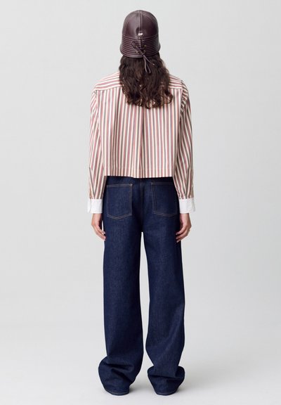Camisa corta a rayas en blanco, rojo y melocotón, combinada con unos pantalones de mezclilla oscuros y sueltos. El modelo lleva un sombrero de cuero marrón con una cinta.