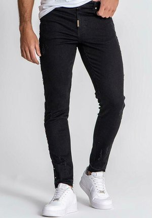 Black denim jeans with a slim fit, featuring distressed details and side button accents, paired with white sneakers.