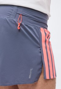 Nahaufnahme einer Person, die blaue Sportshorts mit korallenfarbenen Seitenstreifen, einer korallenfarbenen Kordel und einem ClimaCool-Etikett am Oberschenkel trägt.