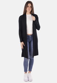 Cardigan lungo nero realizzato in un tessuto morbido con un design aperto davanti. Il modello indossa un top bianco e jeans blu, abbinati a sneakers bianche.