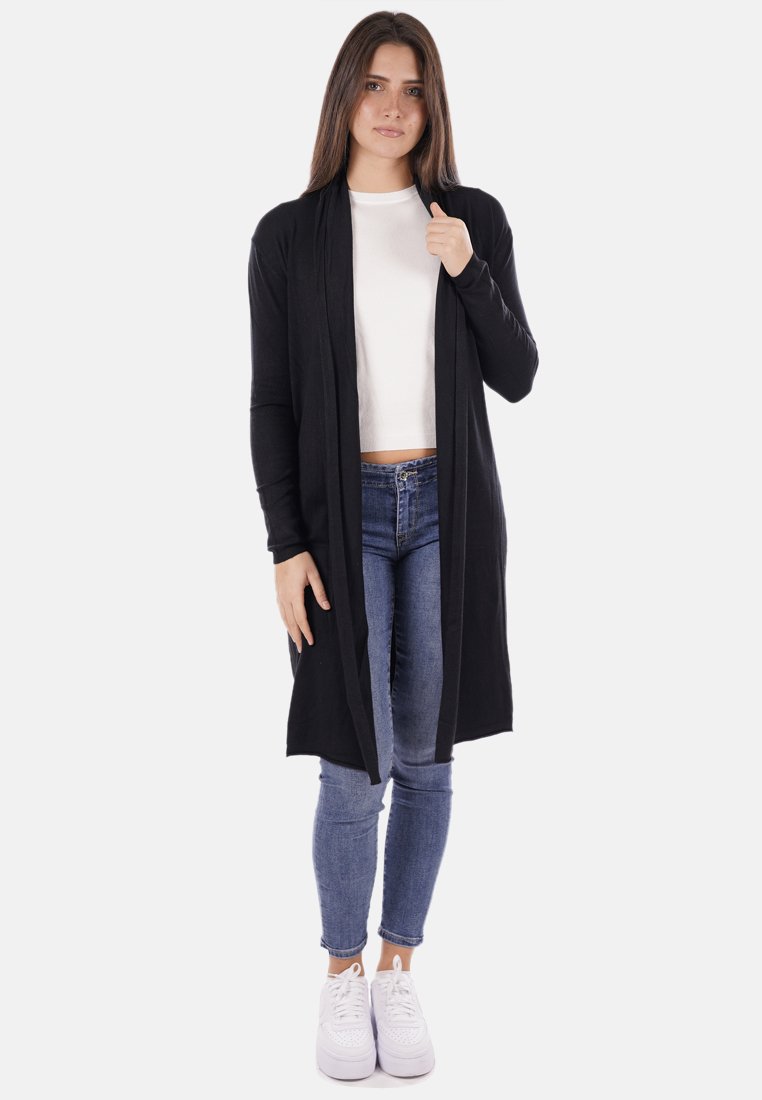 Cardigan lungo nero realizzato in un tessuto morbido con un design aperto davanti. Il modello indossa un top bianco e jeans blu, abbinati a sneakers bianche.