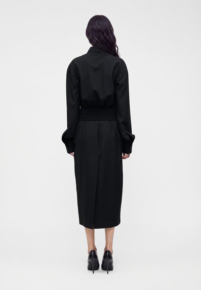Femme aux longs cheveux foncés portant une robe midi noire à manches longues et des chaussures noires à talons hauts, debout de dos sur un fond uni.