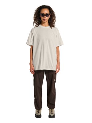 Vrouw met krullend haar, gekleed in zwarte zonnebril, oversized beige t-shirt, donkerbruine broek en beige wandelschoenen, staand met het gezicht naar voren.