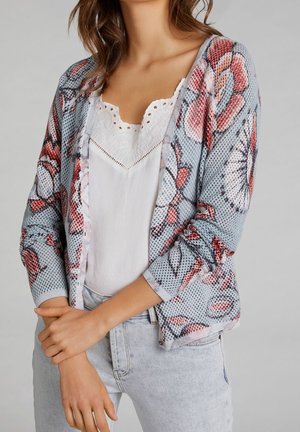 Frau trägt ein weißes Top mit Spitzenbesatz, kombiniert mit hellblauen Jeans und einer floralen, netzgestrickten Strickjacke. Arme verschränkt.
