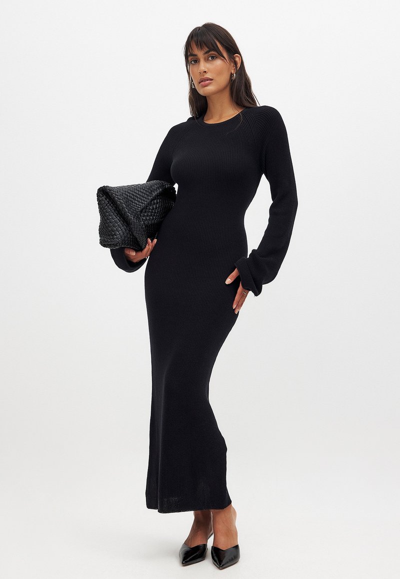 Robe noire en tricot côtelé à manches longues, à coupe ajustée, avec un col rond et un ourlet évasé. Associée à une pochette noire texturée et des chaussures à talons pointus.