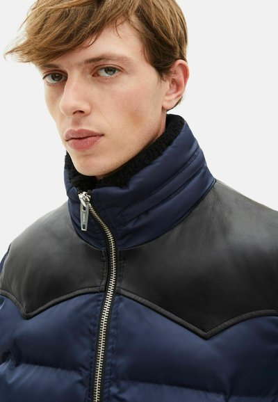 Steppjacke aus marineblauem Stoff mit schwarzen Lederakzenten auf den Schultern. Sie verfügt über einen hohen Kragen, einen silbernen Reißverschluss und ein strukturiertes Futter.