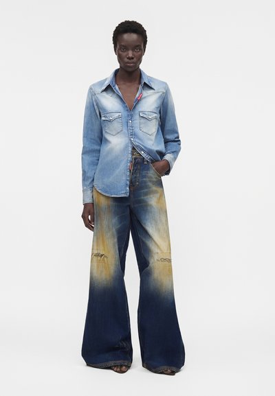 Άτομο που φοράει ξεθωριασμένο ανοιχτό μπλε τζιν πουκάμισο και φαρδιά μπλε τζιν με κίτρινα σημάδια από λευκόνισμα και φθαρμένα γόνατα.