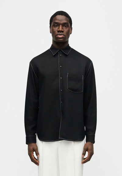 Άνδρας που φοράει μαύρο πουκάμισο με μακριά μανίκια και κουμπιά, με λευκά ραφτά και τσέπη στο στήθος, συνδυασμένο με λευκό παντελόνι, στέκεται όρθιος.