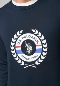 Marineblaue Langarmshirt mit einem weißen Logo mit einem Polo-Spieler, Lorbeerkranz-Akzenten und einem rot-blauen gestreiften Detail.