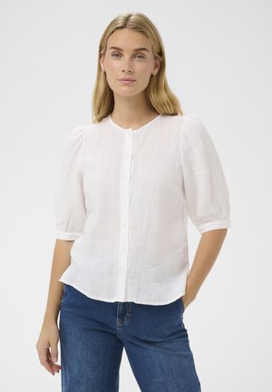 Woman with blonde hair wearing a white button-up blouse with puff sleeves and blue jeans, posing with one hand in pocket against a plain background.