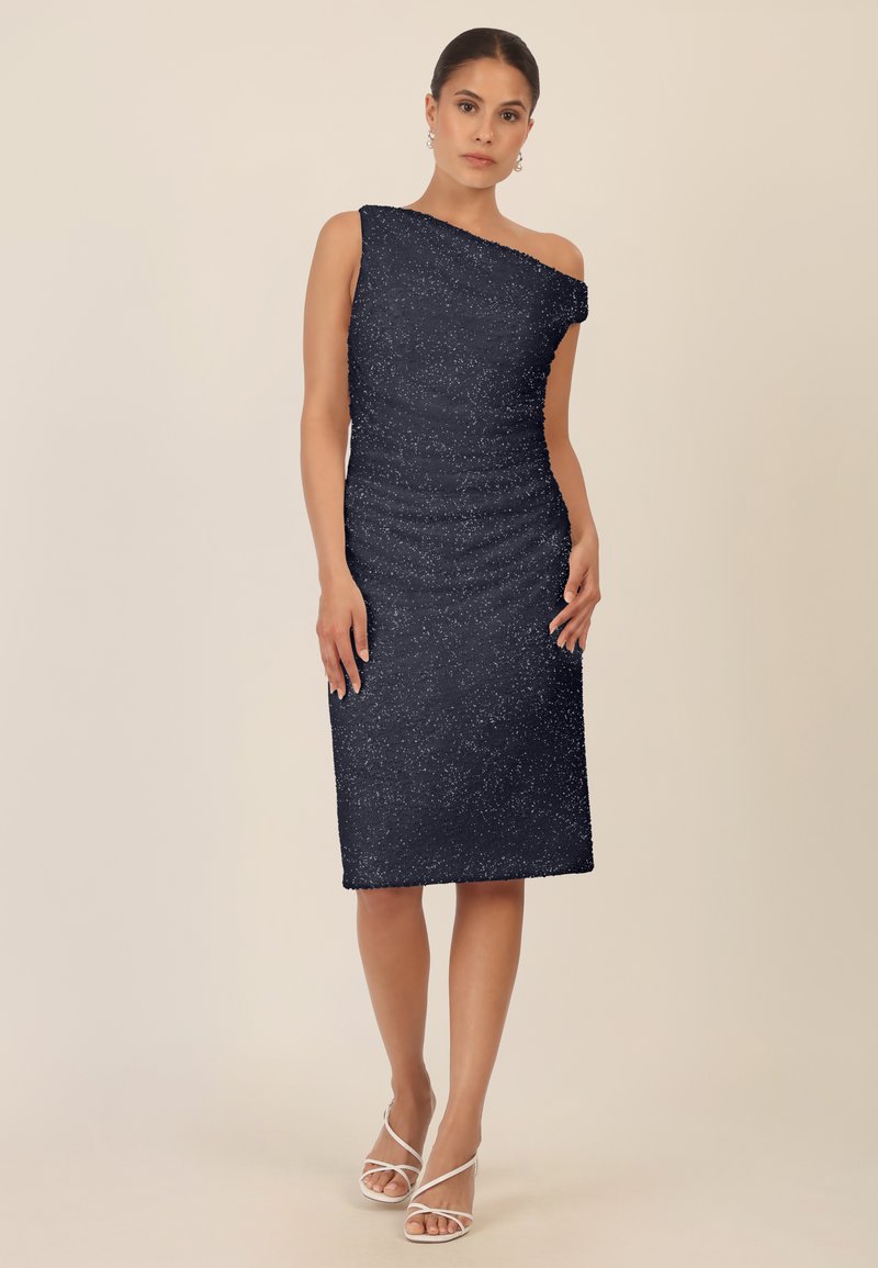 Sukienka midi z jednym ramieniem w kolorze granatowym, o dopasowanym kroju, z teksturowanej tkaniny i subtelnego połysku. Połączona z białymi sandałami na obcasie.