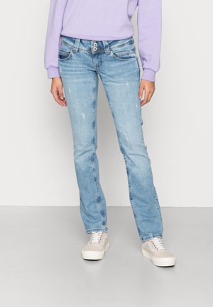 Mujer vestida con jeans ajustados de color azul claro, zapatillas beige y blancas, y una blusa de manga larga lila contra un fondo simple.