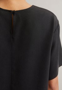 Top nero a maniche corte con scollatura arrotondata e chiusura a oblò sul retro. Il tessuto presenta una sottile trama e un aspetto liscio.