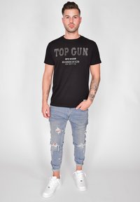 Svart bomull T-shirt med vit "TOP GUN" tryck och text nedanför. Bärs med ljusblå slitna jeans och vita sneakers.