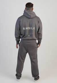 Szürke pamut kapucnis felső, nagy fehér "SIKSILK" felirattal a hátsó részen, hozzá illő szürke nadrággal, laza fazonú, bordás mandzsettákkal, fehér cipővel párosítva.