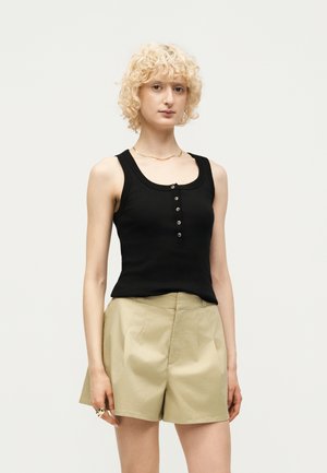 Personne blonde aux cheveux bouclés porte un haut noir sans manches avec des boutons et un short beige taille haute, debout devant un fond uni.