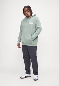 Lichtgroene hoodie met een zak aan de voorkant en trekkoorden. Bevat witte logotekst. Gecombineerd met donkere cargobroeken en sneakers.