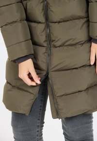Cappotto puffer verde oliva con chiusura a zip, caratterizzato da cuciture orizzontali e una texture liscia e lucida. Una mano sta regolando la zip.