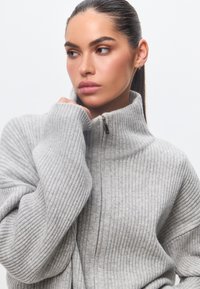Grauer gerippter Reißverschluss-Pullover mit hohem Kragen und eng anliegenden Ärmeln, der über ein strukturiertes Strickmuster verfügt. Einfaches Design mit einem metallischen Reißverschlussteil.
