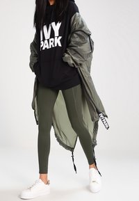 Felpa nera con logo "IVY PARK", giacca bomber verde oliva, leggings verdi oliva, sneakers bianche con dettagli dorati. Tessuti testurizzati, design sportivo.