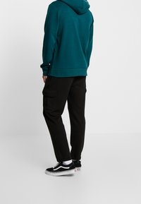 Felpa teal con cappuccio ampio, abbinata a pantaloni cargo neri con tasche laterali. Scarpe nere con suole bianche e calze.