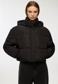 Czarna puchowa kurtka z wysokim kołnierzem i kapturem, o kwadratowym kroju, elastycznymi mankietami i pikowanym wzorze. Gładka, błyszcząca faktura.