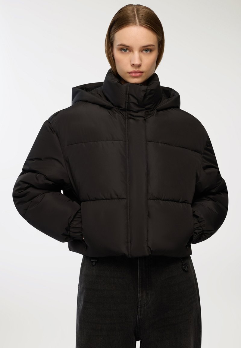 Czarna puchowa kurtka z wysokim kołnierzem i kapturem, o kwadratowym kroju, elastycznymi mankietami i pikowanym wzorze. Gładka, błyszcząca faktura.