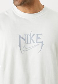 Hvid bomulds t-shirt med et lys gråt Nike-logo design, der har stiliseret skrift og en buet swoosh. Glat tekstur, rund hals.